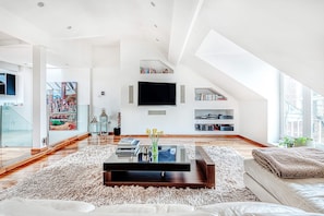 Ein geräumiges Wohnzimmer mit hoher Decke, einem großen Flachbildfernseher an der Wand, einem Holztisch und einem weichen Teppich.