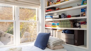 Een slaapkamer met een groot raam, een bed met witte lakens, een blauw kussen en een boekenkast vol boeken en decoratieve spullen.
