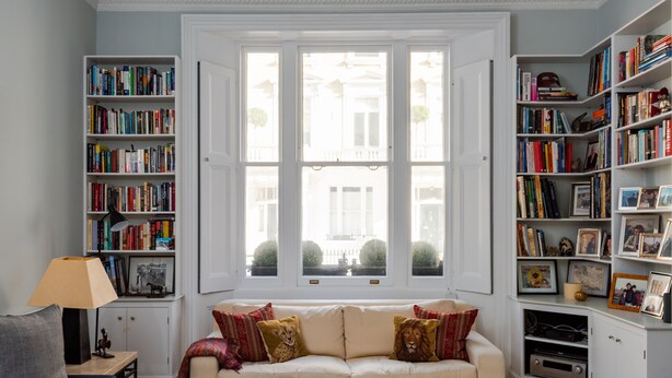 Een gezellige woonkamer met een beige bank, een kleine tafel met een lamp en boekenkasten vol boeken.
