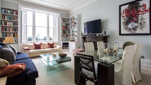 Een woonkamer met een bank, een glazen salontafel, een open haard, een televisie en een boekenkast.
