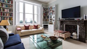 Een woonkamer met een bank, fauteuil, salontafel, open haard, boekenkasten en een televisie.