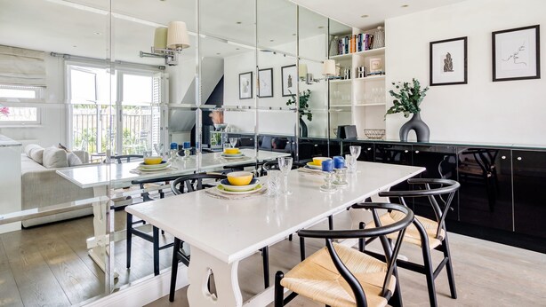 A modern kitchen with a large island, white cabinetry, and a dining area with a white table and black chairs. - The Luxury Treehouse - One bed Little Venice - by Out of Office Lifestyle (London)