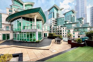 A modern hotel with a curved glass facade, a spacious outdoor area with a seating area, and a view of surrounding high-rise buildings. - The English Rose Luxury 3 bed With Private Terrace - by Out of Office Lifestyle (London)
