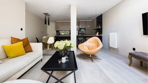 A modern living room with a sofa, coffee table, and a leather chair. There is a kitchen in the background with appliances and a dining area. - The English Gentleman Hampstead Heath - by Out of Office Lifestyle (London)