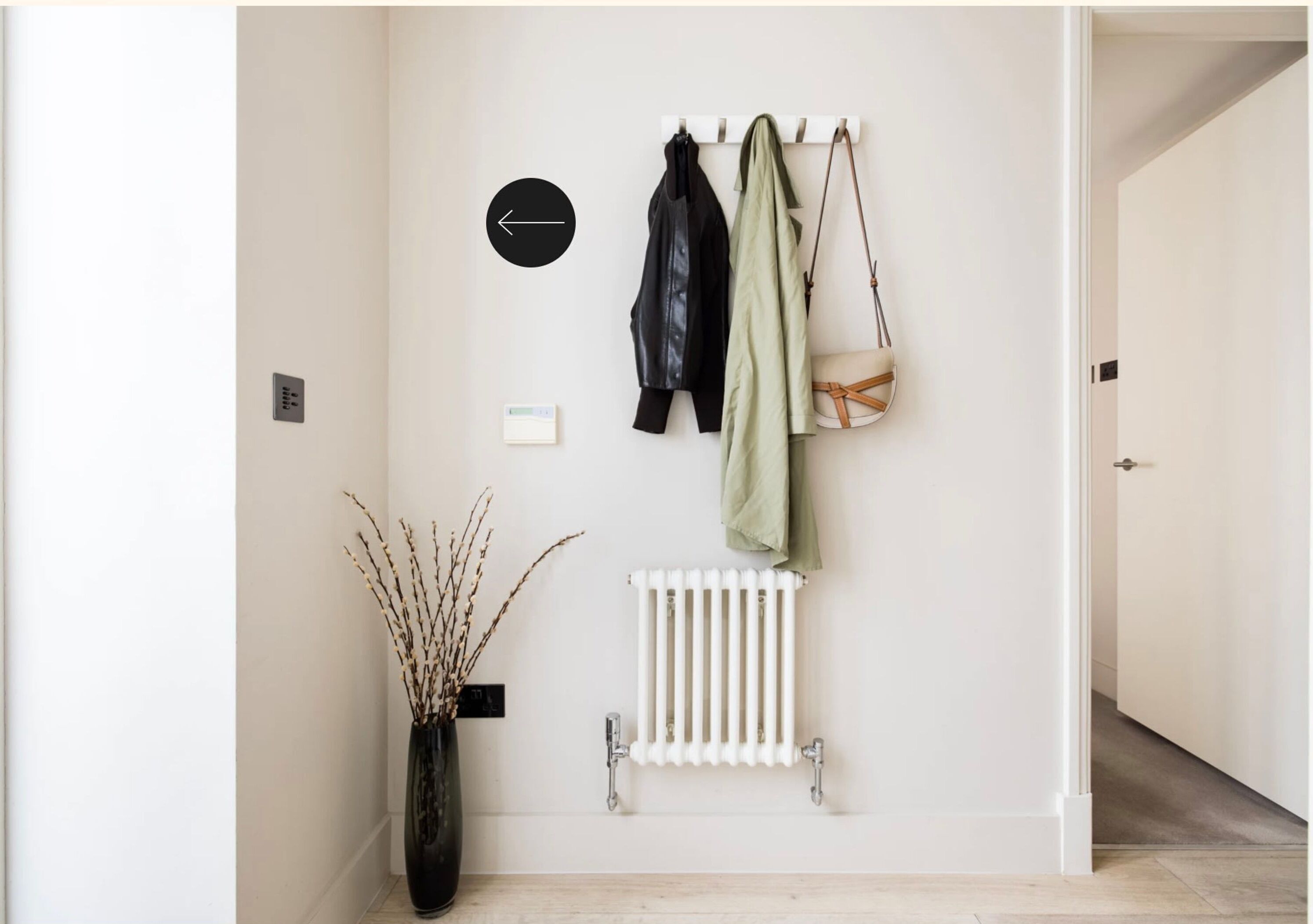 A hallway with a coat rack holding a black jacket, a green jacket, and a beige bag. There is a radiator below the rack and a vase with branches to the left.