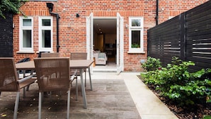 A brick house with a patio area featuring a table and chairs, a black fence, and a garden. - The Bohemian Rhapsody- Garden Apartment in Hampstead - by Out of Office Lifestyle (London)