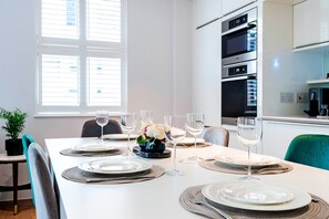 Un coin repas avec une table blanche dressée pour quatre personnes, comprenant des assiettes, des couverts et des verres à vin. Un pot de fleurs se trouve à gauche, et une fenêtre avec des stores est visible à l’arrière-plan.