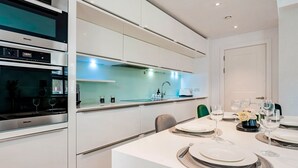 A modern kitchen with built-in appliances, a dining area with a white table set for six, and a view of the adjacent living space. - The Smooth Velvet - by Out of Office Lifestyle (London)