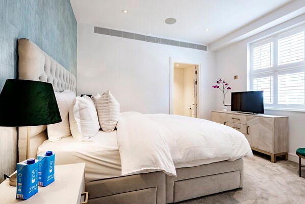 A modern bedroom with a large bed, bedside tables, a TV, and a window with blinds.