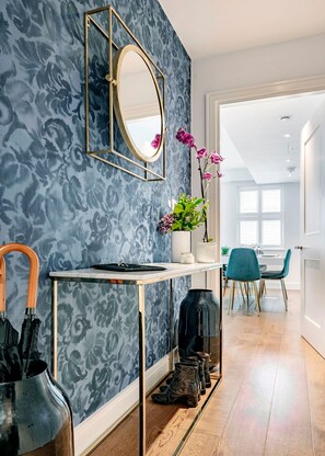 Une chambre moderne avec un mur bleu à motifs, un miroir rond, un vase avec des fleurs, une table avec un plateau, et une paire de bottes.