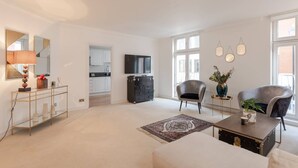 A spacious living room with a sofa, a coffee table, and a television. There is a kitchen area with cabinets and a refrigerator. - The Mayfair Muse - by Out of Office Lifestyle (London)