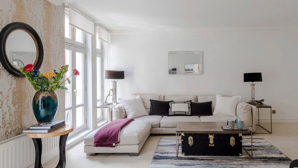 Een moderne woonkamer met een witte bank, een bijzettafeltje en een decoratieve vaas met bloemen. Er hangt een grote spiegel aan de muur en een raam zorgt voor natuurlijk licht.