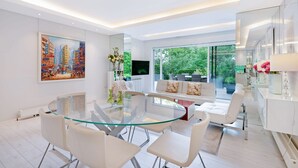 A modern living room with a glass dining table, white sofas, and a large window offering a view of greenery. - The White Peace Garden 3 bed Hampstead - by Out of Office Lifestyle (London)