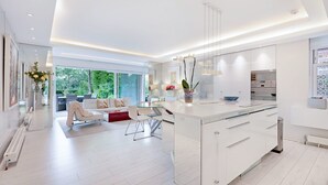 A modern kitchen with white cabinetry, a large island, and a dining area with a glass table and white chairs. - The White Peace Garden 3 bed Hampstead - by Out of Office Lifestyle (London)
