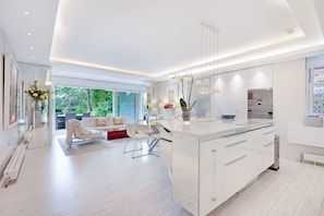 A modern kitchen with white cabinetry, a large island, and a dining area with a glass table and white chairs. - The White Peace Garden 3 bed Hampstead - by Out of Office Lifestyle (London)