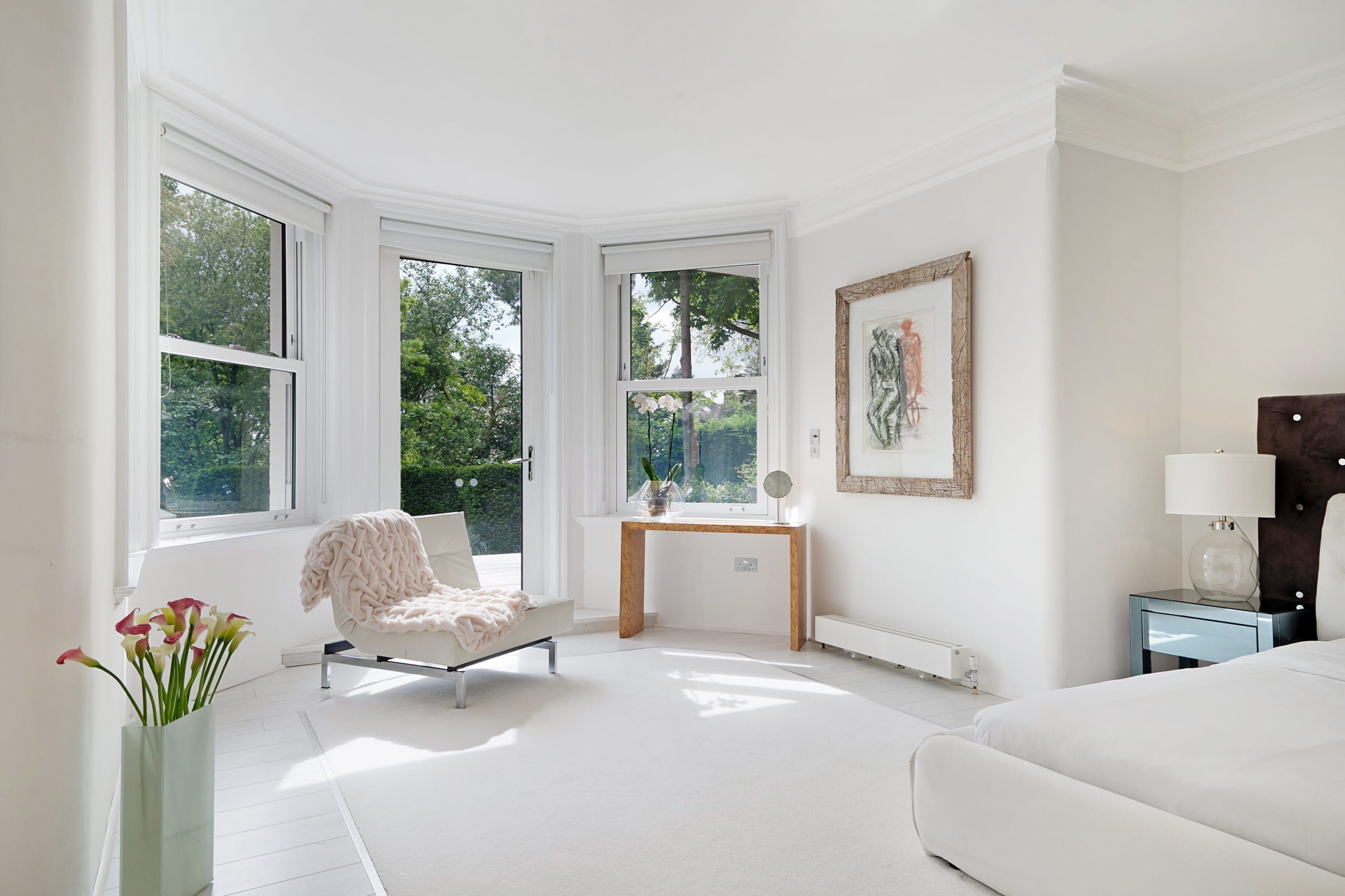 Une pièce lumineuse et blanche, dotée de grandes fenêtres, avec un lit, une chaise, une petite table et une table de chevet.