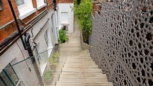 A wooden staircase leading down to a narrow alleyway with a white fence on one side and a brick wall on the other. - The White Peace Garden 3 bed Hampstead - by Out of Office Lifestyle (London)