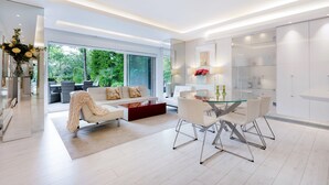 A modern living room with a glass dining table, white sofas, and a large window offering a view of greenery. - The White Peace Garden 3 bed Hampstead - by Out of Office Lifestyle (London)