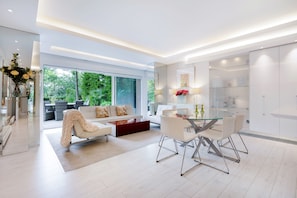 A modern living room with a glass dining table, white sofas, and a large window offering a view of greenery. - The White Peace Garden 3 bed Hampstead - by Out of Office Lifestyle (London)