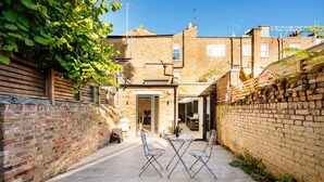 A narrow courtyard with a brick wall on one side, a wooden fence on the other, and a small table with chairs in the center. - The Blooming Minimalist- by Out of Office Lifestyle (London)