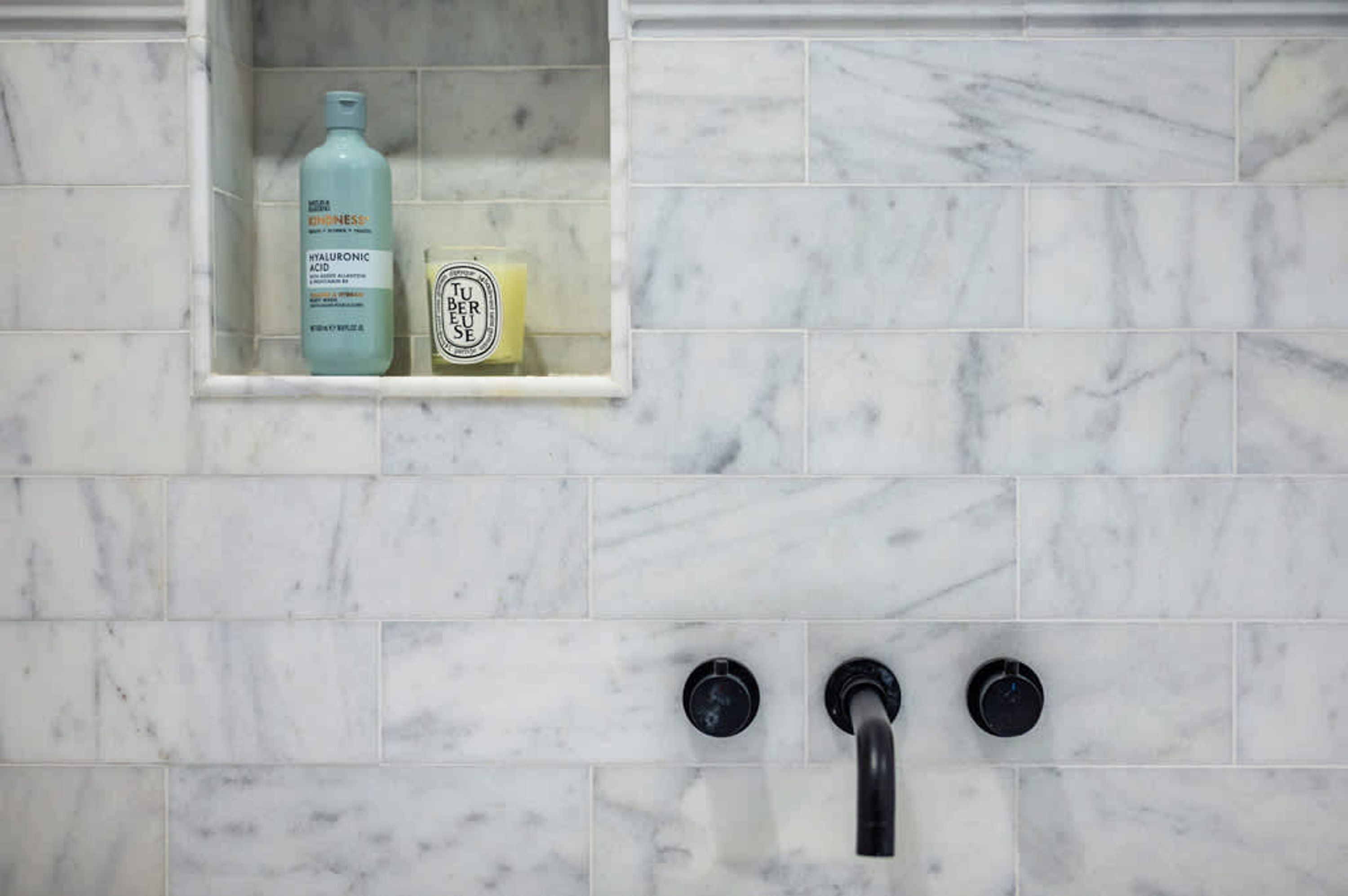 A bathroom with a marble tiled wall, a shelf with toiletries, and a black faucet.