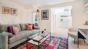 Un salon moderne avec un canapé gris, une table basse en verre et un tapis à motifs floraux rouges. Au fond, on aperçoit une cuisine blanche avec des meubles blancs et un luminaire suspendu.