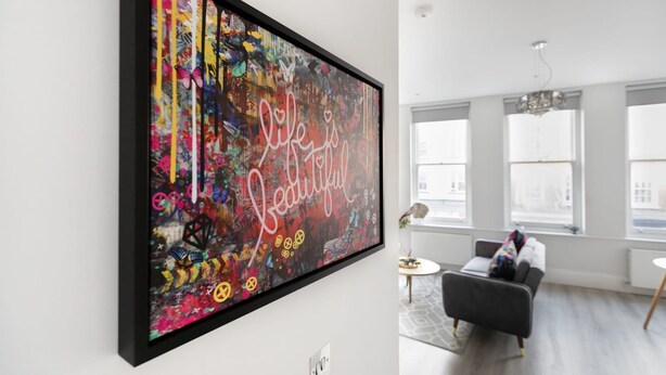 Un salon moderne avec un grand tableau abstrait au mur, un canapé gris, une petite table ronde et de grandes fenêtres laissant entrer la lumière naturelle.