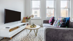 Un salon moderne doté d’un téléviseur à écran plat, d’un meuble de rangement blanc, d’un canapé gris agrémenté de coussins colorés, d’une table basse ronde et de grandes fenêtres offrant une vue sur la ville.