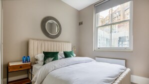Une chambre bien rangée, avec un lit, une table de chevet et une fenêtre donnant sur des immeubles.