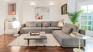 Ein modernes Wohnzimmer mit einer grauen Eckcouch, einem Couchtisch und einer Stehlampe. An der Wand hängt ein Bild und auf der rechten Seite steht ein Blumentopf.