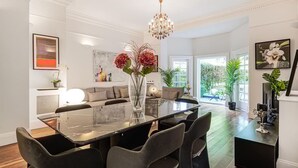 Ein stilvoller Essbereich mit einem Glastisch, schwarzen Stühlen und einer Blumenvase. Über dem Tisch hängt eine Chandelier, im Hintergrund ist eine Couch zu sehen.