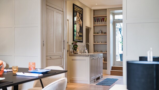 A modern kitchen with white cabinetry, a marble countertop, and a potted plant. There is a dining area with a table and chairs, and a black lamp on a side table.
