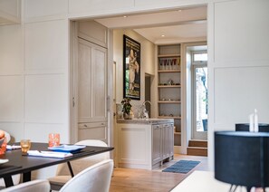 A modern kitchen with white cabinetry, a marble countertop, and a potted plant. There is a dining area with a table and chairs, and a black lamp on a side table. - The Notting Hill Townhouse - by Out of Office Lifestyle (London)