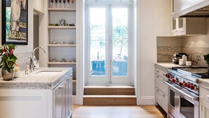 A modern kitchen with white cabinetry, a marble countertop, a built-in oven, and a large window overlooking a garden.