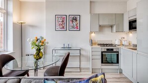 A modern kitchen with white cabinets, a gas stove, and an oven. There is a dining area with a glass table and chairs, and two framed artworks on the wall.