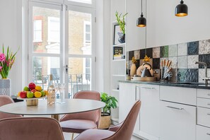 A modern kitchen with white cabinets, a black countertop, and a dining area with a round table and pink chairs. - Lady on the Lane- 2 bed Primrose Hill - by Out of Office Lifestyle (London)