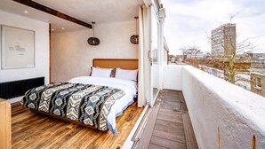 A bedroom with a wooden floor, a bed with patterned bedding, a headboard, and a view of the city through large windows. - Long Term Stay- Notting Hill by Out of Office Lifestyle (London)