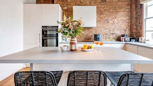 A kitchen with a large island, white cabinets, a brick wall, and a hanging light fixture. - Long Term Stay- Notting Hill by Out of Office Lifestyle (London)