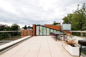 A rooftop terrace with a glass wall, outdoor seating, and a view of the surrounding greenery. - The Modern Glasshouse - by Out of Office Lifestyle (London)