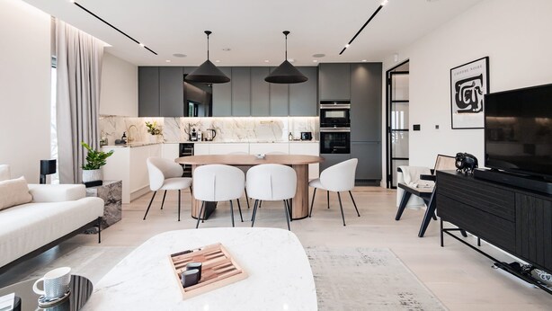 A modern living room with a sofa, coffee table, and a TV. The room has a dining area with a table and chairs. There is a kitchen in the background with a stove and cabinets.