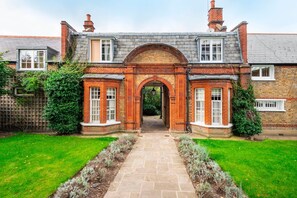 A traditional brick house with a central archway, a stone pathway leading to the entrance, and a well-maintained garden. - The Kensington Gatehouse - by Out of Office Lifestyle (London)