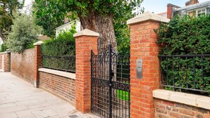 A gated entrance with a brick wall and metal gate, a tree, and a hedge. - The Kensington Gatehouse - by Out of Office Lifestyle (London)