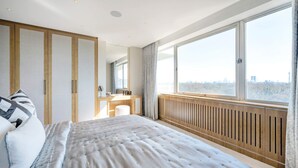 A modern bedroom with a large bed, a vanity with a mirror, and a view of a cityscape through large windows. - Regents Park Penthouse (London)