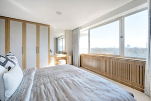 Une chambre moderne avec un grand lit, une coiffeuse avec un miroir et une vue sur le paysage urbain grâce à de grandes fenêtres.