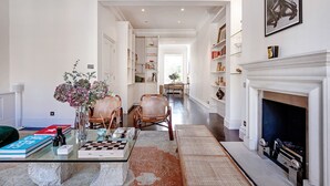 Un salón amplio con chimenea, una mesa de centro con libros y un jarrón, y una estantería llena de libros.