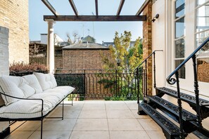 Eine überdachte Terrasse mit einem weißen Sofa, einem schwarzen schmiedeeisernen Geländer und einer Treppe zu einer weißen Tür.