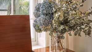 Un aparador de estilo antiguo de madera con un tocadiscos incorporado, un florero con hortensias azules y eucalipto, y vista de un edificio a través de una ventana.