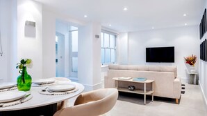 A modern living room with a sofa, a TV mounted on the wall, and a dining area with a table set for two. - The Urban Minimalist- Notting Hill 2 Bed (London)