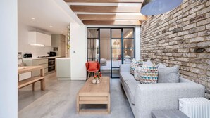 Un interior moderno con cocina, comedor y sala de estar, destacando una pared de ladrillo, vigas de madera y un gran ventanal.