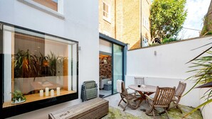 Un patio moderno con una mesa y sillas de madera, un banco y un ventanal con vista a un muro de ladrillo.